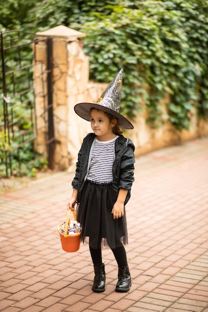 Niña linda disfrazada de bruja sosteniendo un cubo de calabaza jack-o-lantern con dulces y dulces. Truco o trato para niños en las vacaciones de Halloween.