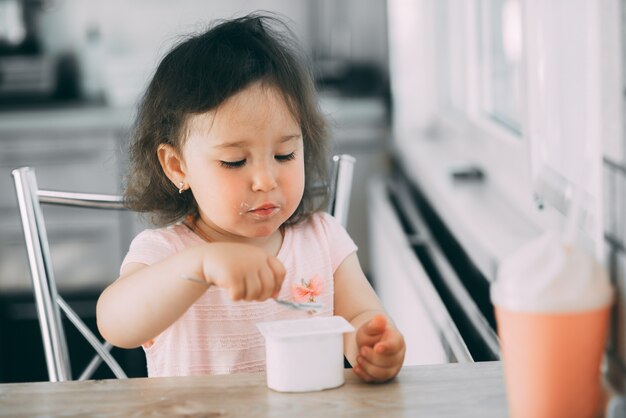 Niña linda y divertida comiendo yogur en la cocina con un vestido rosa por la tarde linda