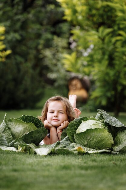 Niña linda se divierte y está acostado sobre la hierba verde cerca de las coles.