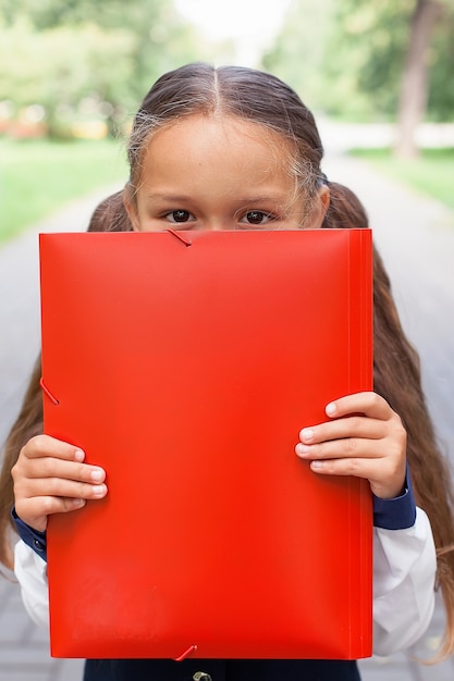 Una niña linda con dos colas sostiene una carpeta con papeles en sus manos. Caminando por el parque, volviendo a la escuela