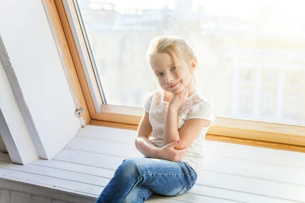 Niña linda dulce sonriente en jeans y camiseta blanca sentada en el alféizar de la ventana en la sala de estar de luz brillante en casa y pensando Infancia, escolares, juventud, concepto de relajación