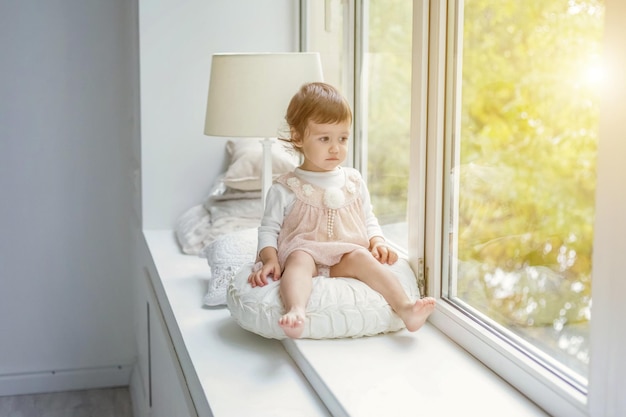 Niña linda dulce sonriente en vestido rosa pastel de pie en el alféizar de la ventana en la luminosa sala de estar en casa y sonriendo. Infancia, preescolar, juventud, concepto de relax