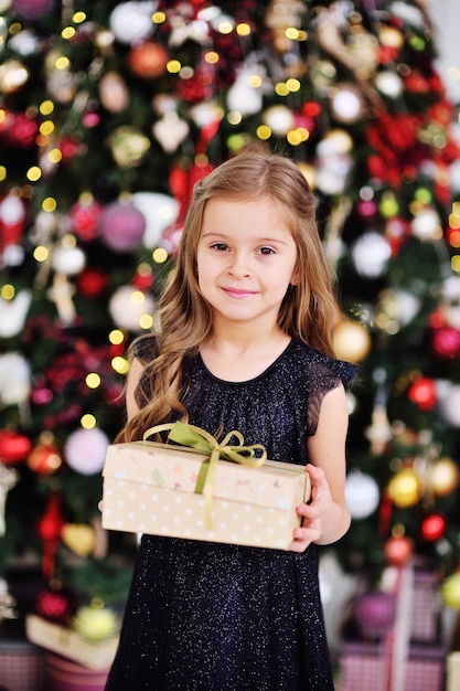 Una niña linda con un elegante vestido negro sosteniendo una caja con un regalo y sonriendo en el fondo del árbol de Navidad y la decoración navideña.