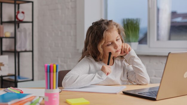 Niña linda de la escuela primaria que estudia en casa usando la computadora portátil. La colegiala tiene una lección en línea. Educación a distancia, educación en el hogar