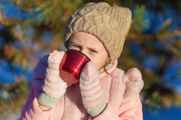 Niña linda está bebiendo de una taza en el bosque de invierno. Infancia feliz. Niños al aire libre Concepto de vacaciones divertidas de invierno