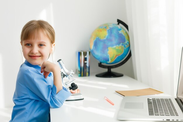 Niña linda está sentada a la mesa con su computadora portátil y estudiando en línea