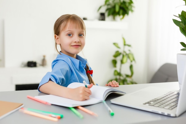 Niña linda está sentada a la mesa con su computadora portátil y estudiando en línea