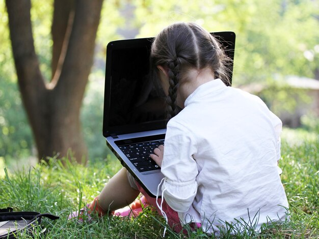 Una niña linda está sentada en el parque con su computadora portátil,