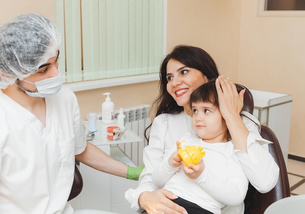 Una niña linda está sentada en el sillón dental con su madre en la clínica El médico está hablando con el paciente Visitando al dentista con niños