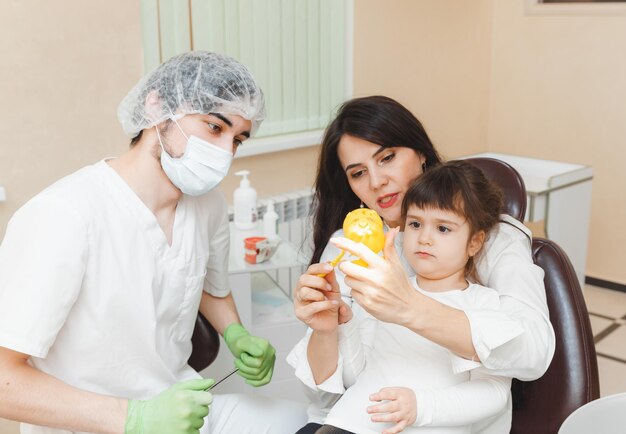 Una niña linda está sentada en el sillón dental con su madre en la clínica El médico está hablando con el paciente Visitando al dentista con niños