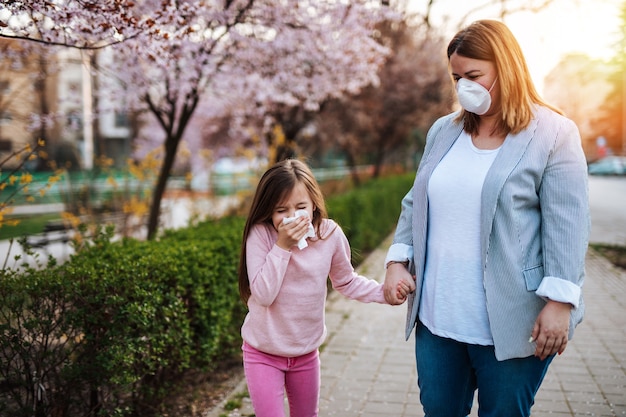 Niña linda estornudando en el parque mientras camina en el parque con su madre. Alergia, gripe, concepto de virus.