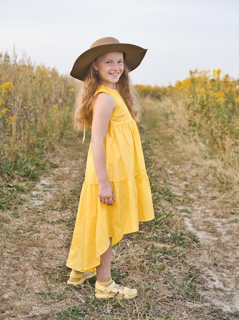 Niña linda feliz y sonriente con vestido amarillo y sombrero camina en un campo Estilo de vida al aire libre de verano