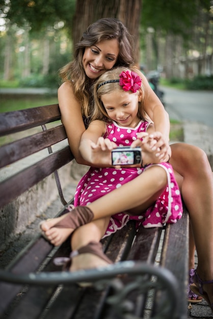 Niña linda feliz y su madre posando para un autorretrato en un banco en el parque.