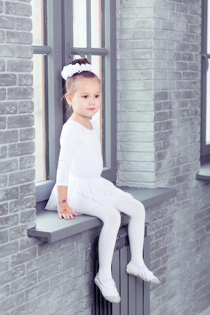 Niña linda en forma de danza blanca sentada en la ventana en el estudio de danza moderna