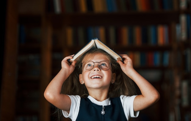 Niña linda con gafas se encuentra en la biblioteca llena de libros