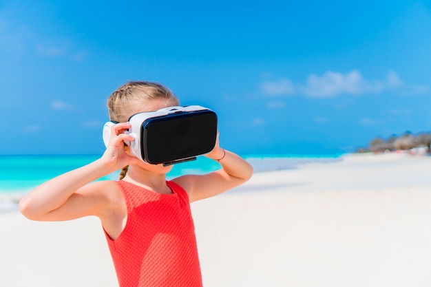 Niña linda con gafas de realidad virtual VR. Adorable niña mira las gafas virtuales en la playa blanca