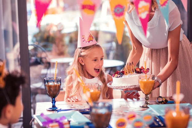 Niña linda con gorro de fiesta mirando su pastel de cumpleaños