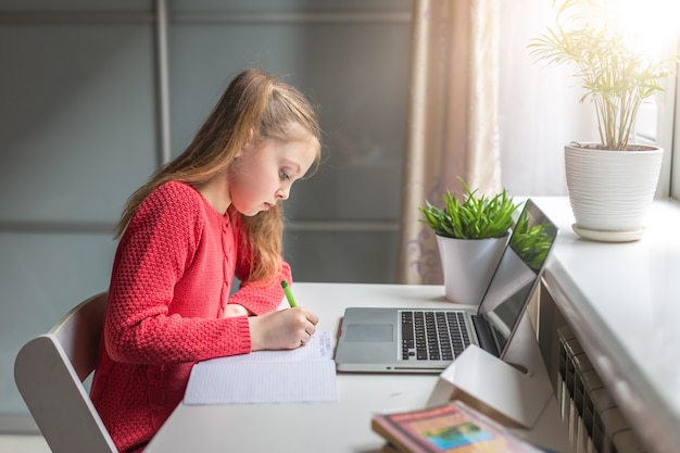 Niña linda haciendo la tarea seriamente usando la computadora portátil en casa para educación en línea, estudio en casa y comunicación en línea. Aprendizaje a distancia para niños. Distancia social. Quédate en casa.