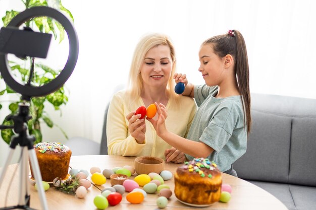 Niña linda y hermosa madre joven sentadas juntas en la mesa de la cocina usando un teléfono inteligente en línea en Pascua.