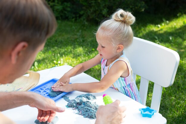 Niña linda jugando con arena cinética junto con el padre al aire libre en el patio trasero en verano