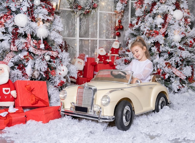 Niña linda jugando con un coche de juguete grande en la habitación decorada de Navidad