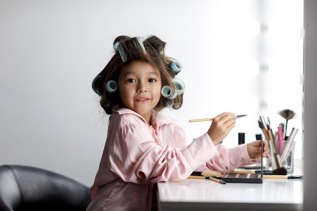 Niña linda jugando con el cosmético de su madre frente al espejo en casa