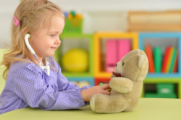 Niña linda jugando a la enfermera inspeccionando el oso de peluche con estetoscopio