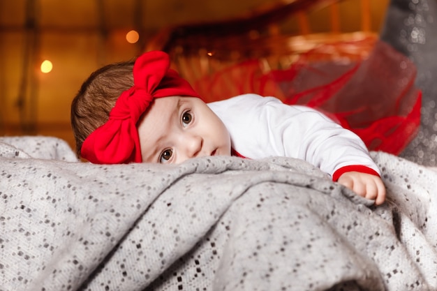 Niña linda en lazo rojo en la cabeza y suéter blanco acostado sobre una manta, un fondo de Navidad en un estudio con un paisaje navideño.