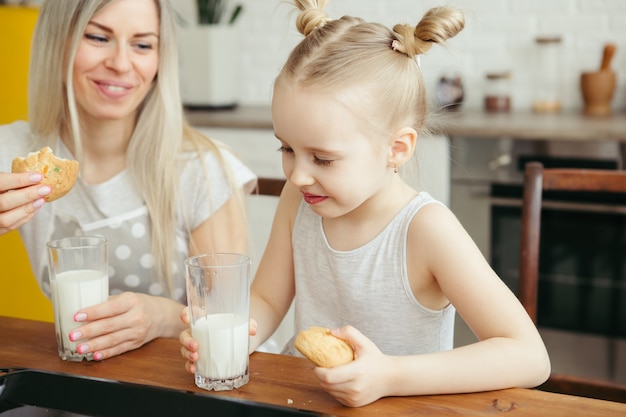 Niña linda y mamá comiendo galletas recién horneadas con leche en la cocina. Familia feliz. Viraje.