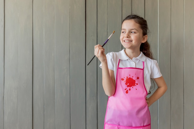 Niña linda con la mano en las caderas que sostienen la brocha que se coloca delante de la pared de madera gris