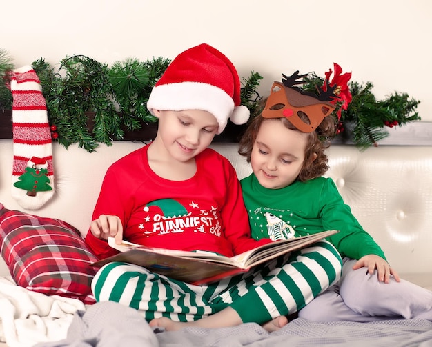 Niña linda con máscara de ciervo divertido y niño con gorro de Papá Noel y pijama de Navidad leyendo un libro.