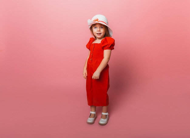 Niña linda con un mono rojo y un sombrero blanco posando sobre un fondo rosa