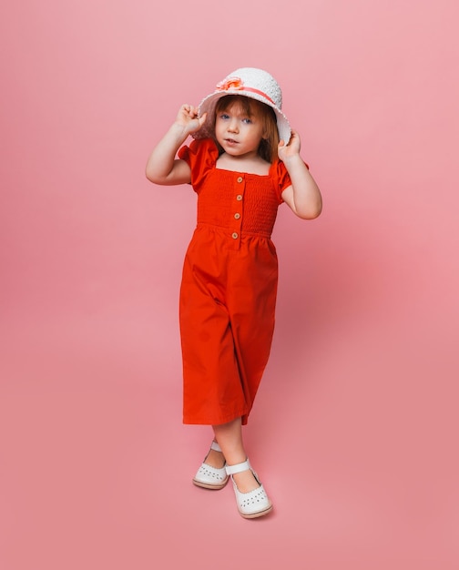 Niña linda con un mono rojo y un sombrero blanco posando sobre un fondo rosa