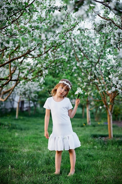 Niña linda del niño 5-6 años que sostiene la flor que se coloca en el jardín de la primavera de la flor, con vestido blanco y corona floral al aire libre, se acerca la temporada de primavera