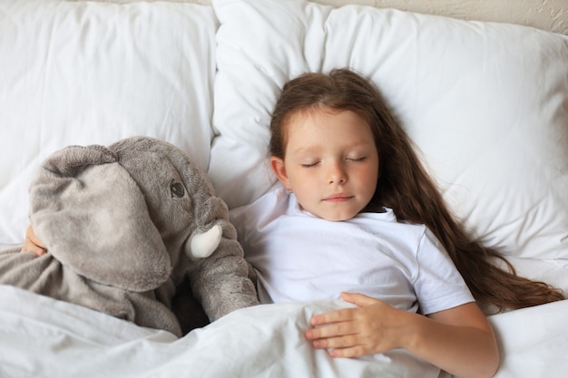 La niña linda del niño duerme en la cama con un elefante de juguete.