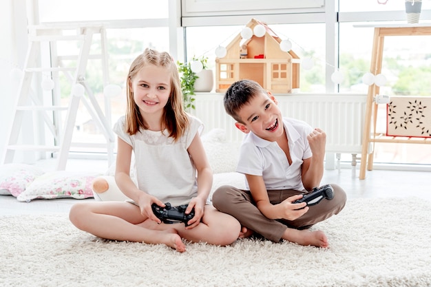 Niña linda y niño con joysticks jugando en la consola en el interior