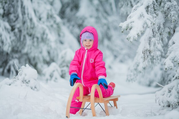 Niña linda de ojos azules en trineo en un bosque nevado. Jardín de invierno. Árboles en la nieve.