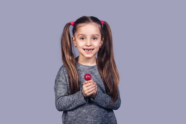 Una niña linda con una paleta roja en sus manos muestra sus dientes estropeados. El concepto de caries dental debido al abuso de dulces. aislado sobre fondo gris