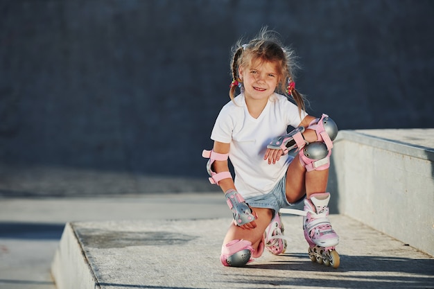 Niña linda con patines al aire libre se sienta en la rampa para deportes extremos.