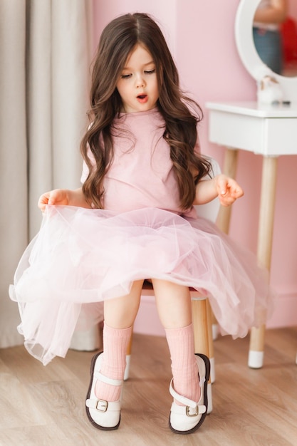 Una niña linda con el pelo largo y un vestido elegante en casa está sentada en una silla en su habitación