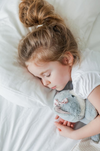 Una niña linda con el pelo rizado duerme en una cama blanca con su juguete de peluche Sueño saludable El niño está descansando en casa