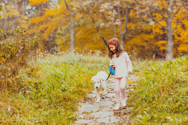 Niña linda y perro golden retriever con una correa caminando en el parque de otoño