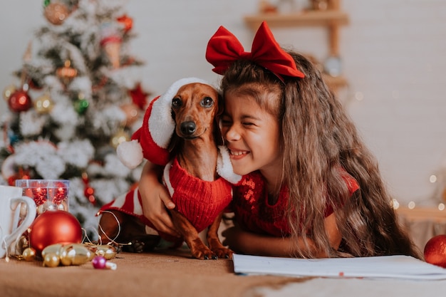 Niña linda y un perro salchicha enano en un traje de Santa Claus celebran la Navidad y el Año Nuevo