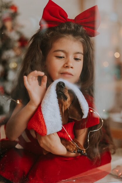 Niña linda con un perro salchicha en un traje de Santa está sentada en la ventana Felices fiestas