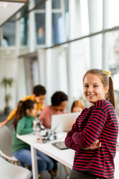 Niña linda de pie frente a un grupo de niños que programan juguetes eléctricos y robots en el aula de robótica