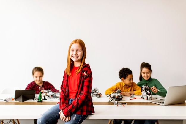 Niña linda de pie frente a un grupo de niños que programan juguetes eléctricos y robots en el aula de robótica