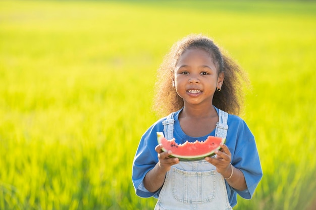 Niña linda de piel negra comiendo sandía al aire libre campo de arroz verde telón de fondo niño africano comiendo sandía