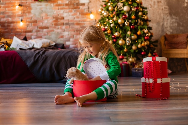 Niña linda en pijama cerca del árbol de Navidad saca del gatito de la caja de regalo