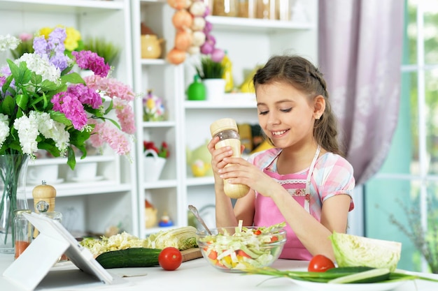 Niña linda preparando ensalada fresca en la mesa de la cocina con tableta en casa