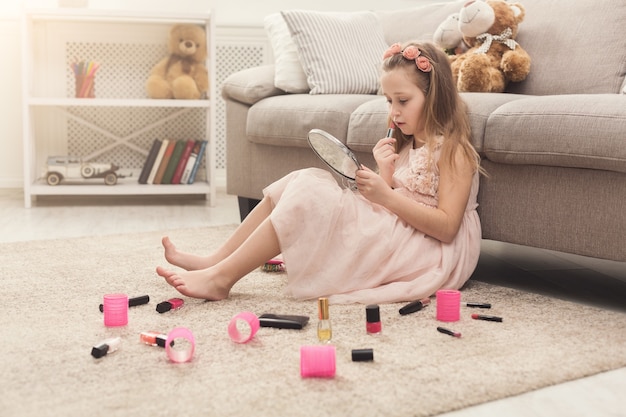 Niña linda probando los cosméticos de su mamá. Bonita niña sentada tiñéndose los labios con lápiz labial, en la alfombra del piso entre una gran cantidad de productos de belleza. Pequeña fashionista haciendo maquillaje, espacio de copia